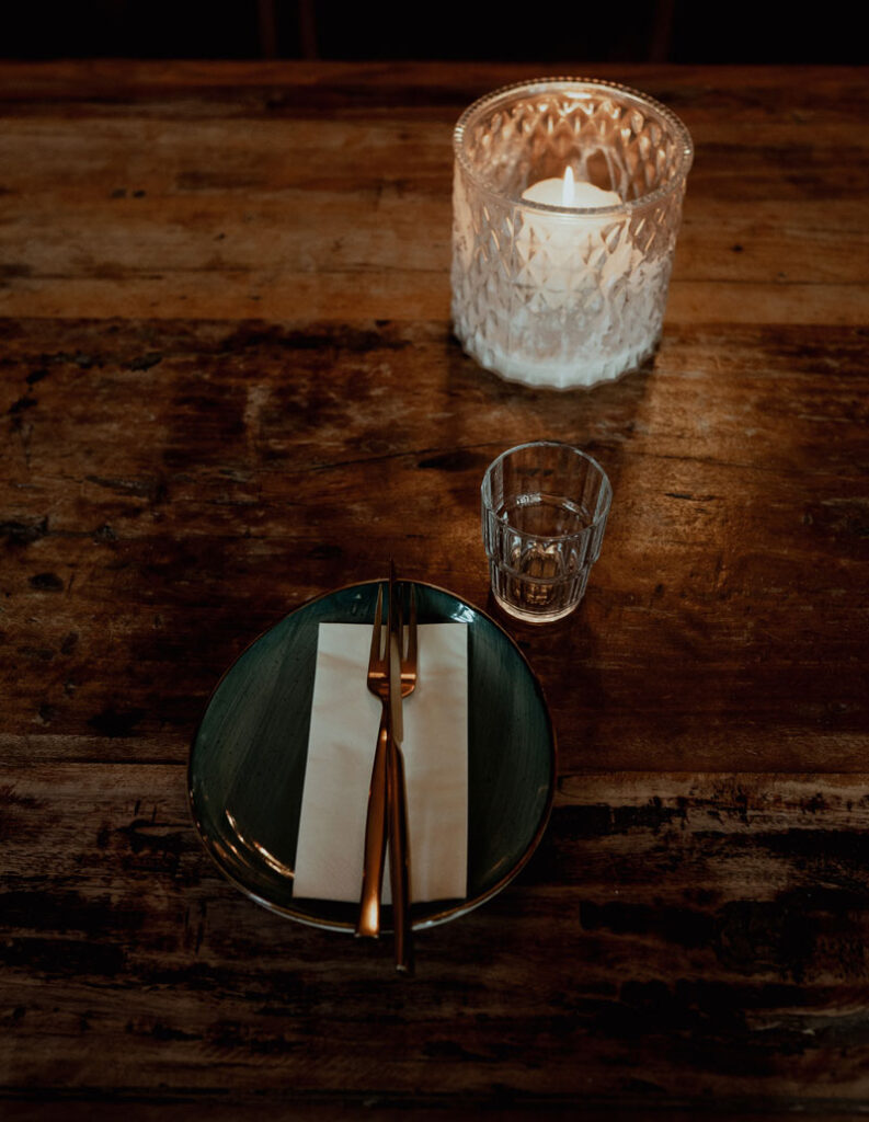 Gedeckter Tisch einer Hochzeit vor dem Essen. Grüner Teller mit Serviette, Messer und Gabel. Daneben steht eine Kerze und ein Wasserglas.