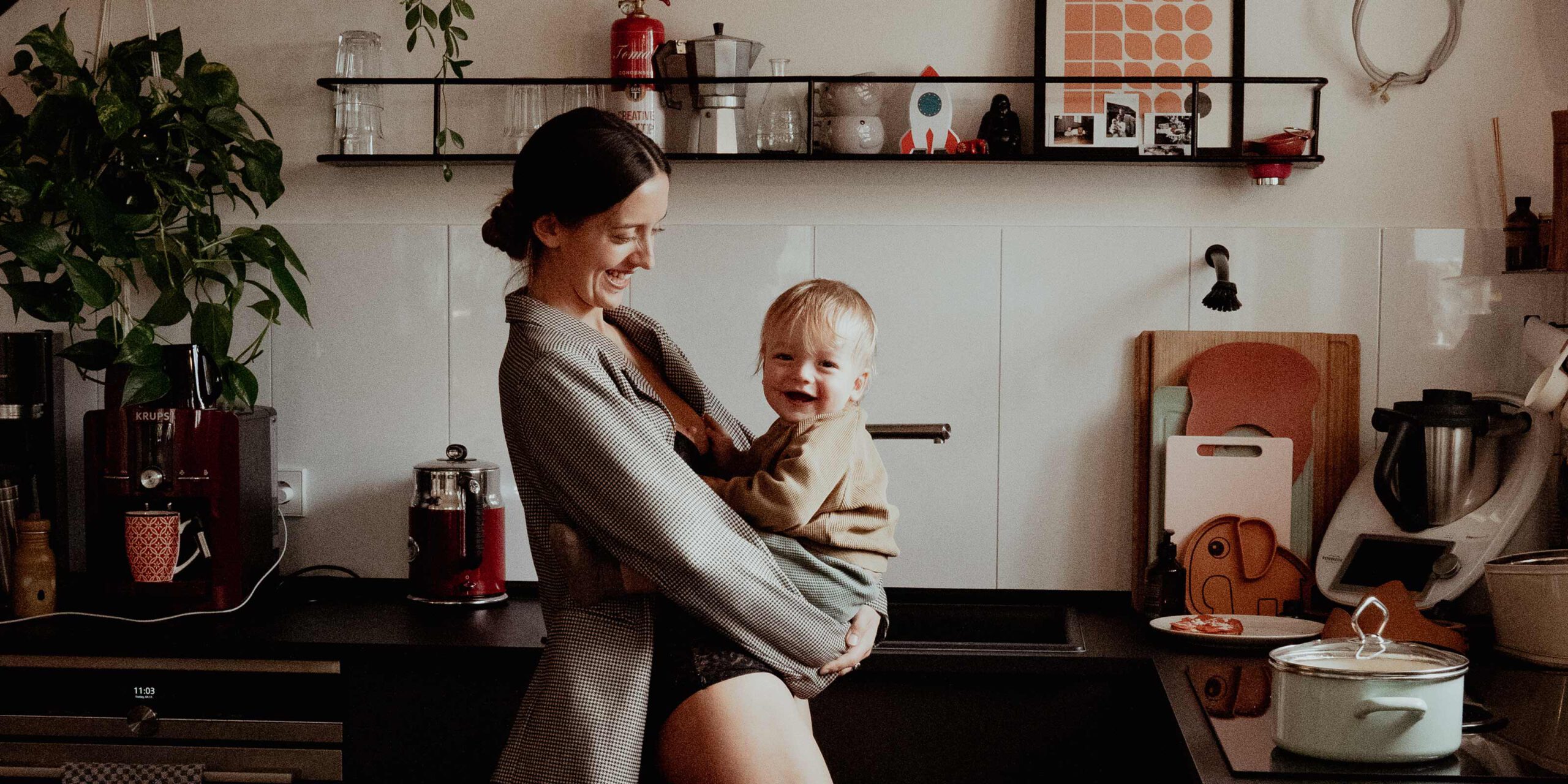 Eine Frau steht in Ihrer Küche mit Ihrem Kind im Arm und lacht. Familienfotografin Alina Lowe